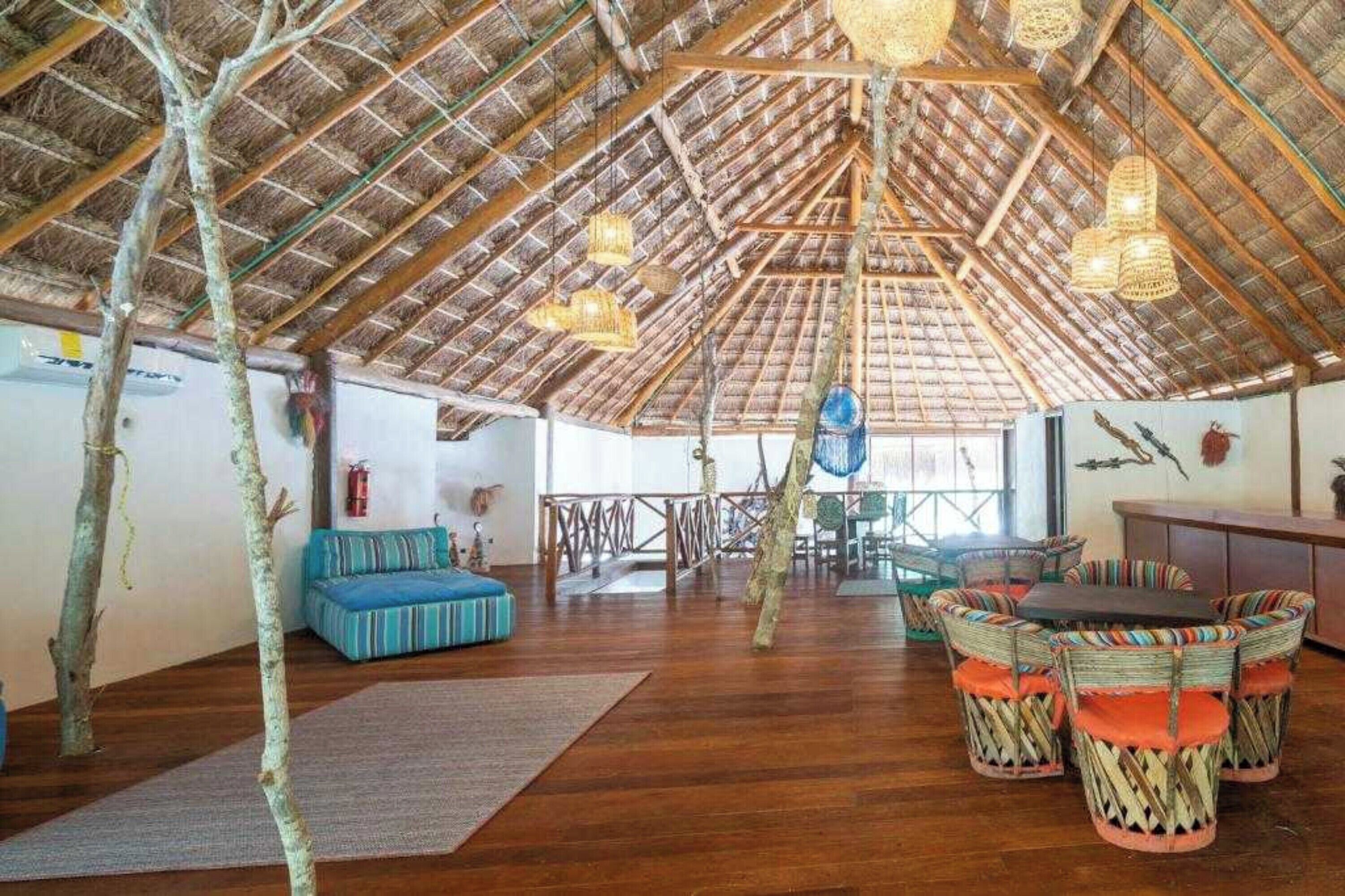 Amplia habitación con techo de paja, suelo de madera y una zona de estar central con sillas tejidas de colores.