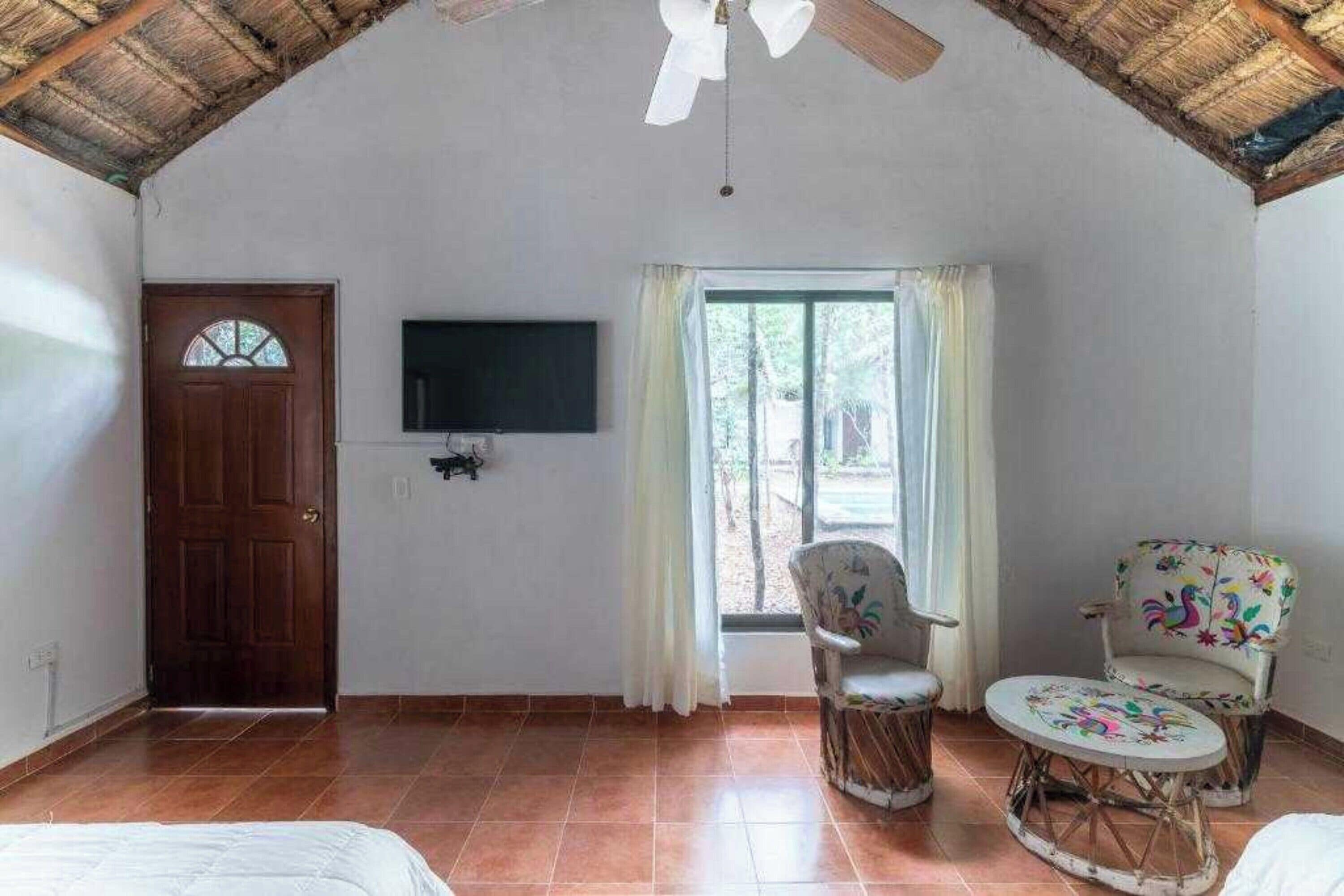 Una habitación con puerta de madera, televisor de pantalla plana, ventana con cortinas y dos sillones de mimbre con una mesita.