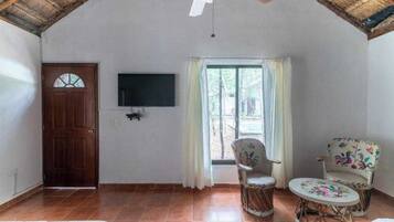 Una habitación con puerta de madera, televisor de pantalla plana, ventana con cortinas y dos sillones de mimbre con una mesita.