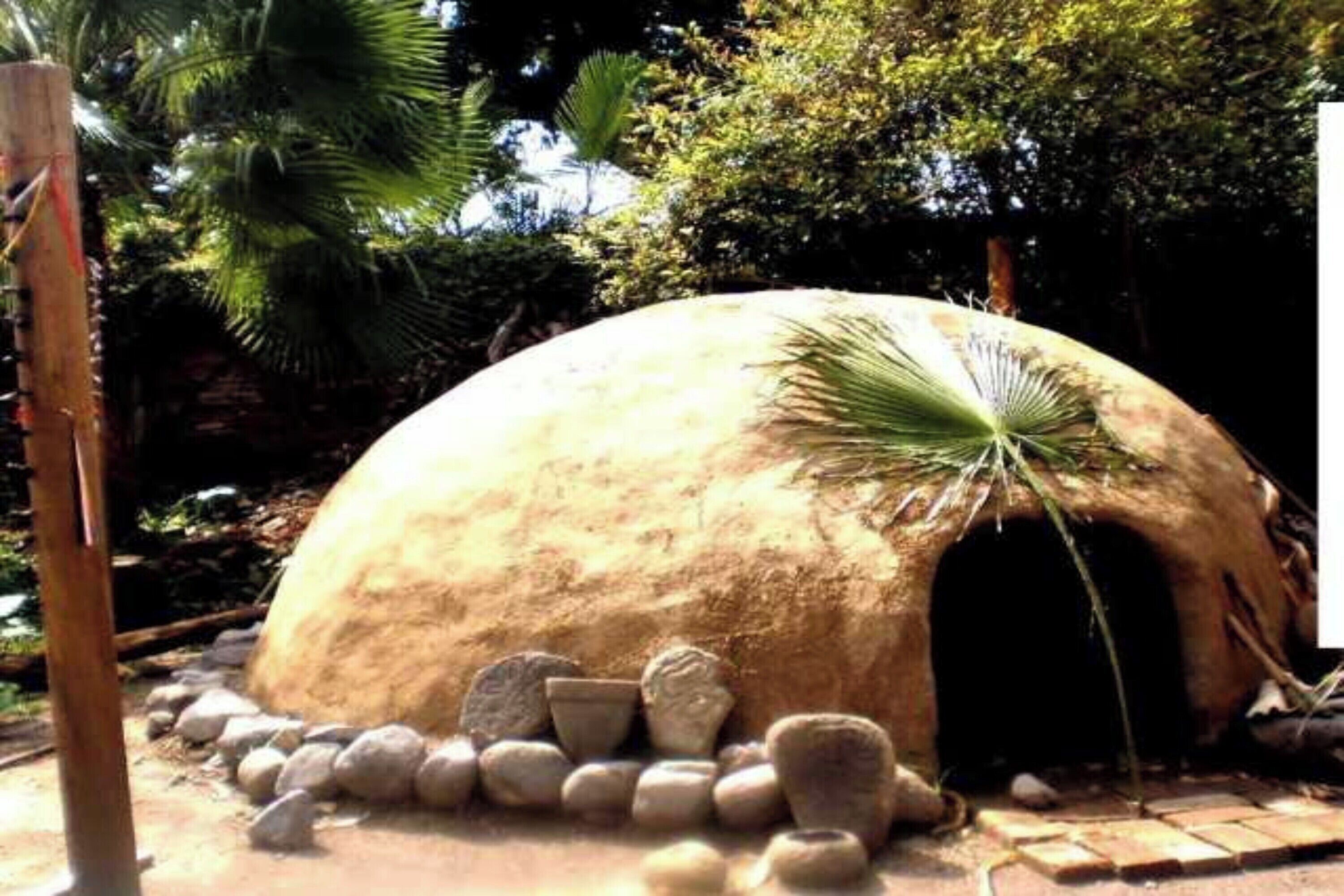 Una estructura abovedada con una hoja de palma en la parte superior, rodeada de rocas y árboles.