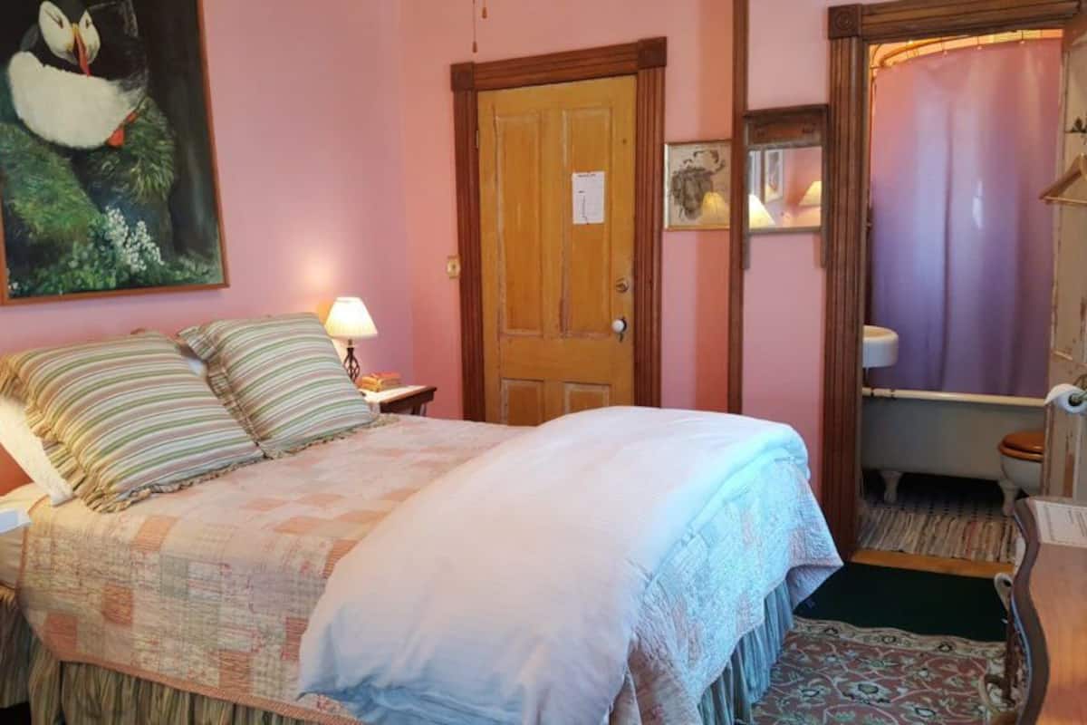 A bedroom with a bed, two bedside tables, a nightstand, a door, a bathroom with a toilet, and a shower curtain.