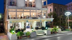 Un hôtel à plusieurs étages, reconnaissable à son grand panneau « PRESTIGE », doté de balcons à chaque étage et d’un hall d’entrée bien éclairé agrémenté de plantes en pot.