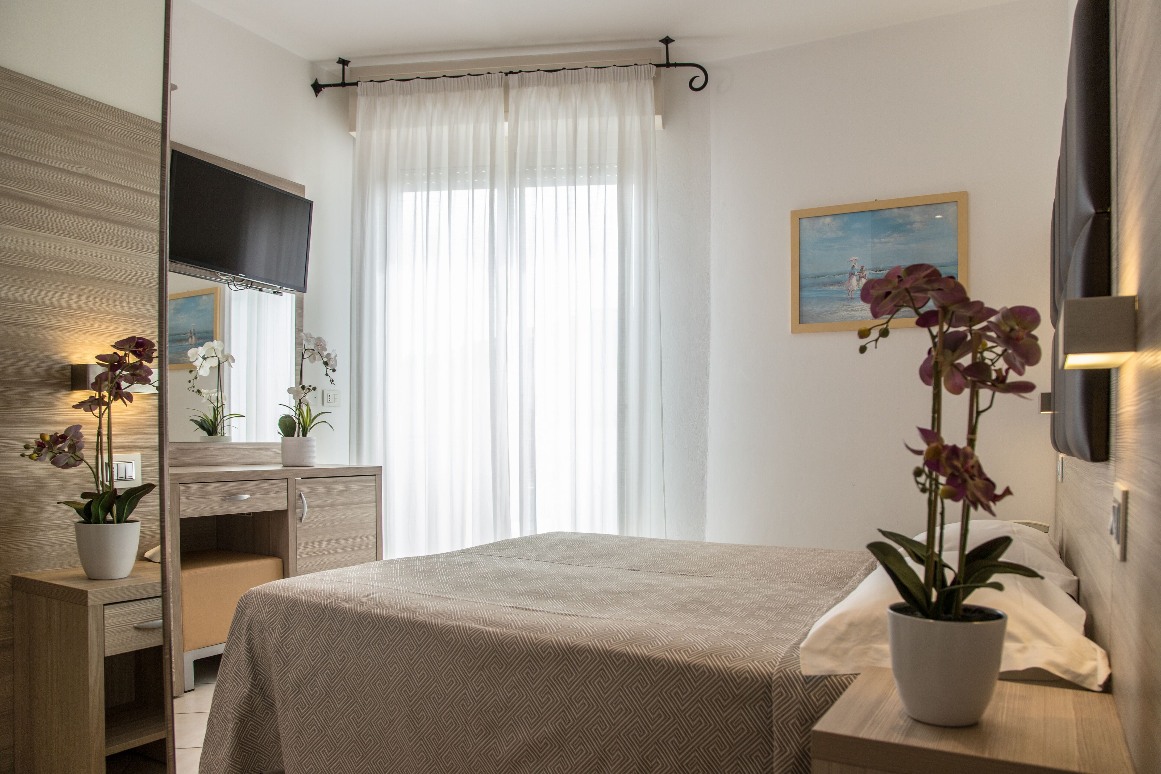 A hotel room with a bed, a bedside table with a potted orchid, a mirror, a flat-screen TV, and a window with sheer curtains.
