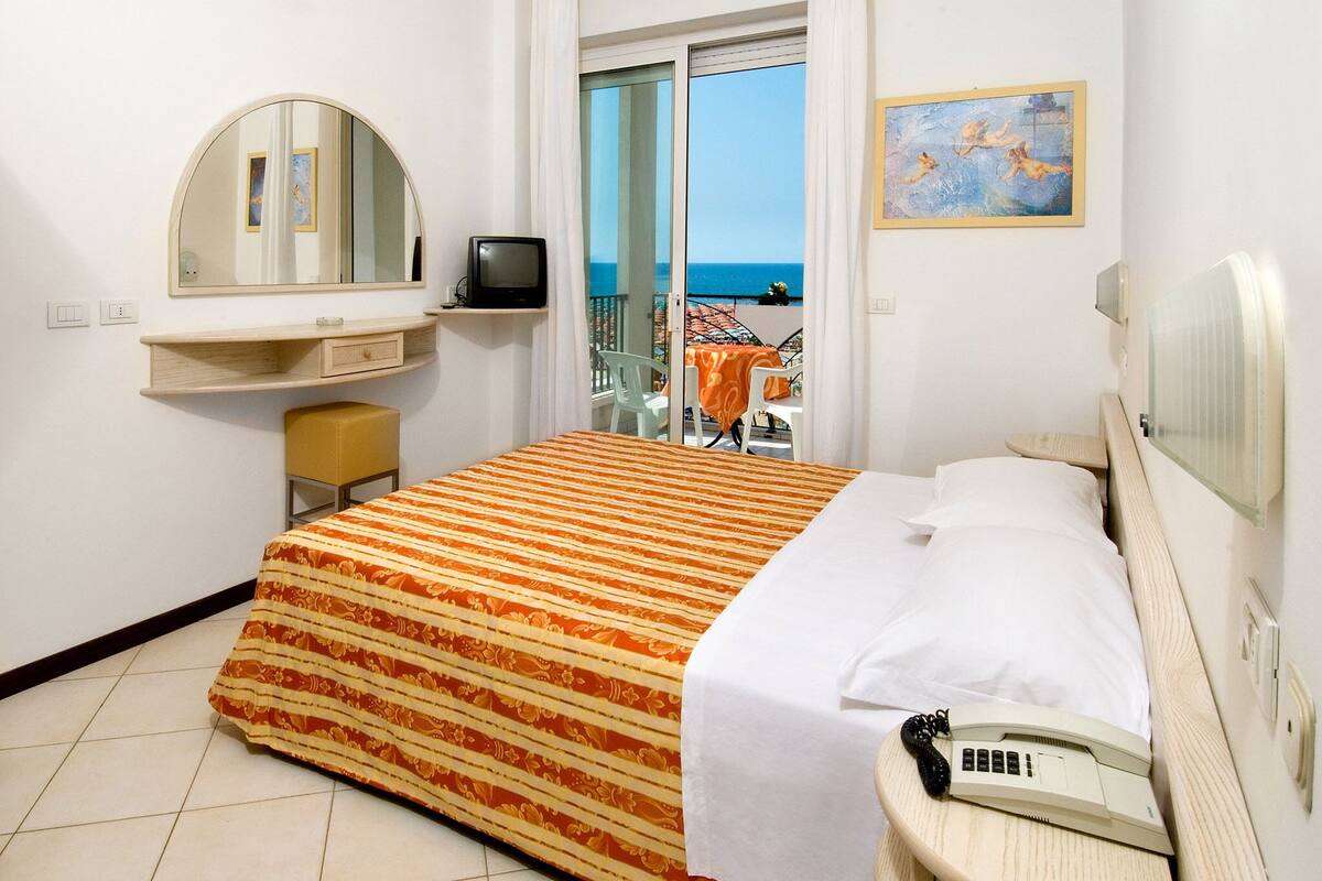 Une chambre d’hôtel équipée d’un lit, d’un téléphone sur la table de chevet, d’une télévision, d’un miroir et offrant une vue sur la mer grâce à une porte vitrée.