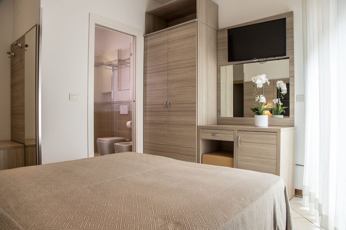 Une chambre moderne avec un grand lit, une armoire, une coiffeuse avec un miroir et un téléviseur à écran plat fixé au-dessus du miroir.
