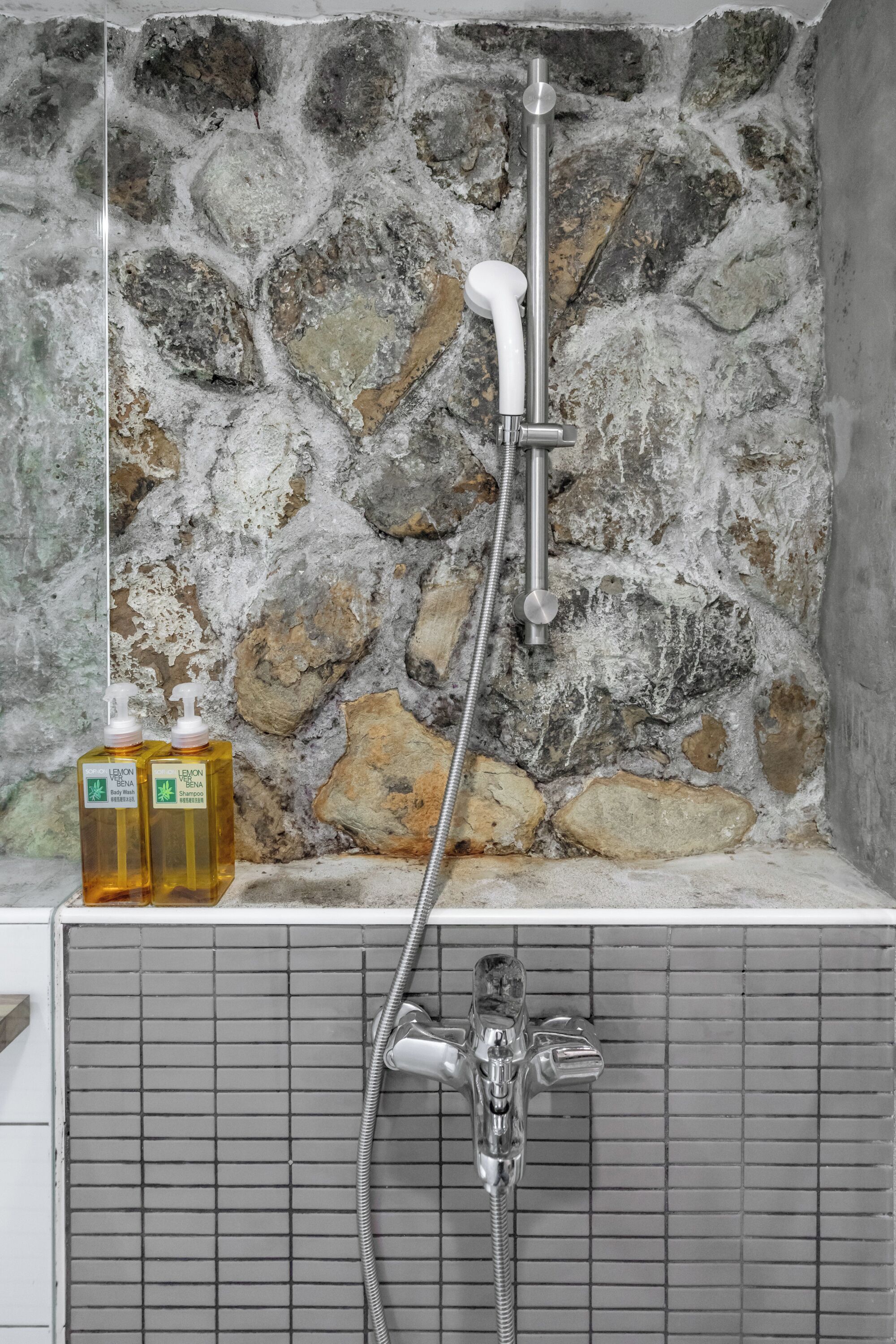 A shower with a stone wall backdrop, a handheld showerhead, and two bottles of liquid soap.