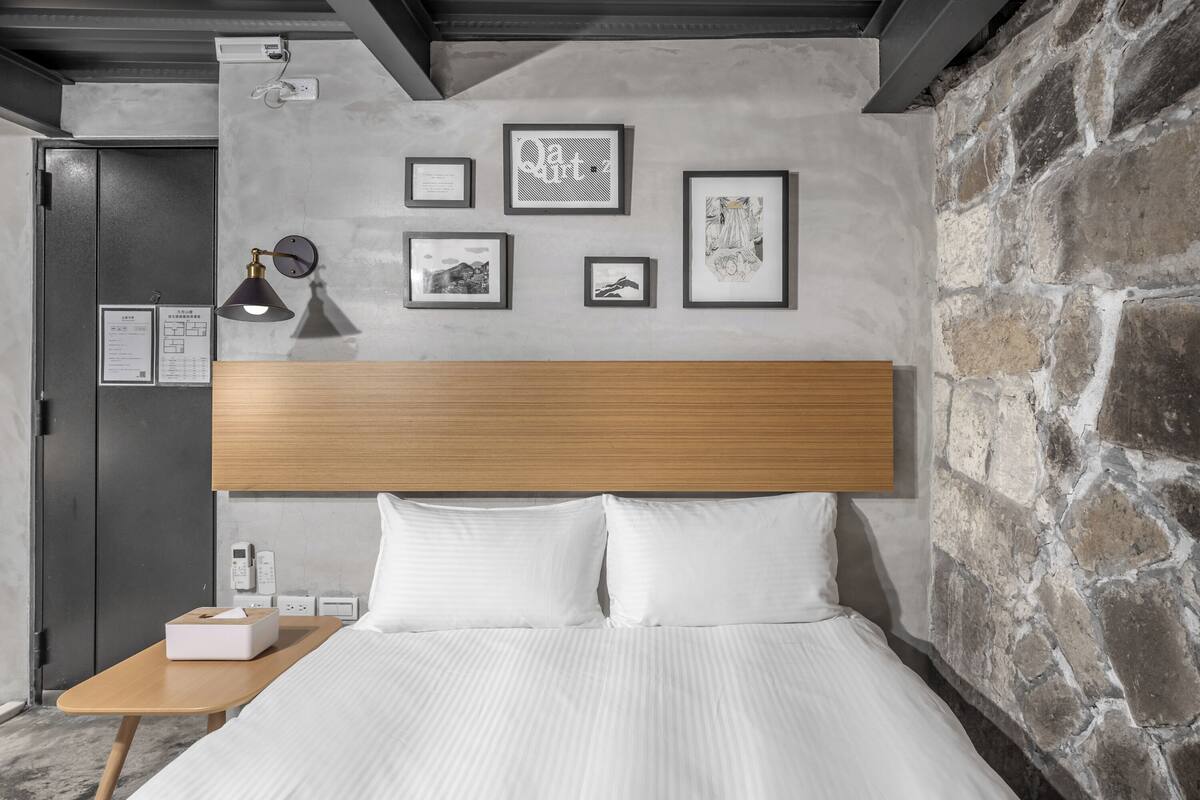 A hotel room with a wooden headboard, white bedding, a bedside table with a lamp, and framed artwork on the wall.