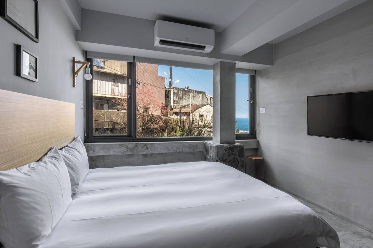 A modern hotel room with a large bed, a flat-screen TV, and a view of the cityscape through large windows.