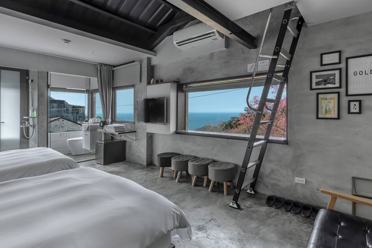 A modern hotel room with a large bed, a desk, a TV, and a view of the sea.