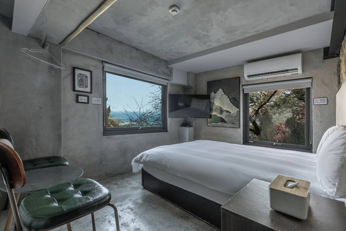 A modern bedroom with a large bed, two windows, a chair, and a bedside table with a tissue box.