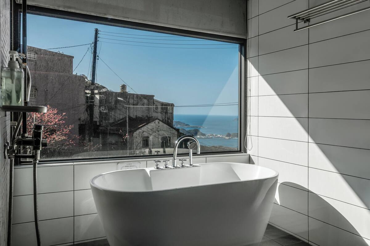 A modern bathroom with a freestanding bathtub, a large window offering a view of buildings and the sea, and a towel rack on the wall.