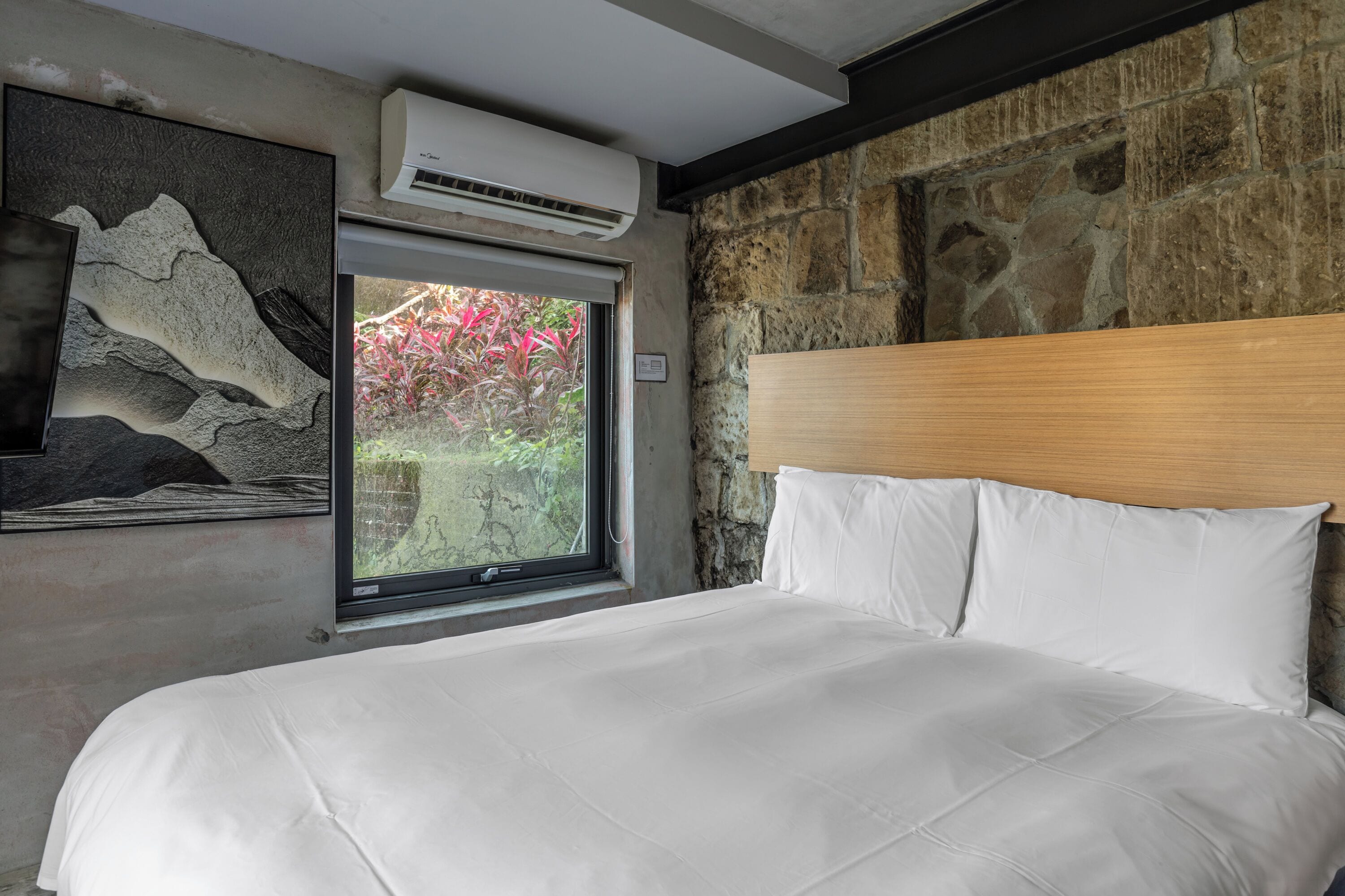 Un dormitorio con pared de piedra, un ventanal grande y una cama con ropa de cama blanca.