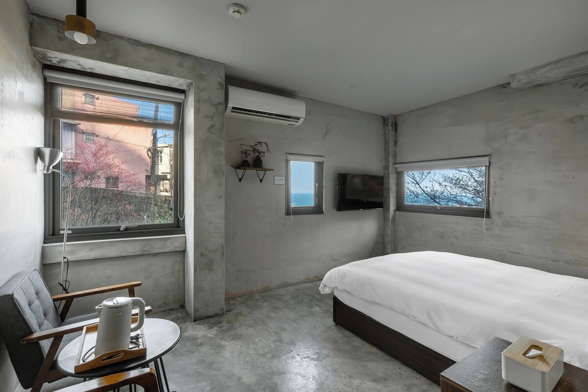 A bedroom with a bed, a desk with a kettle, a chair, a window, and an air conditioning unit.