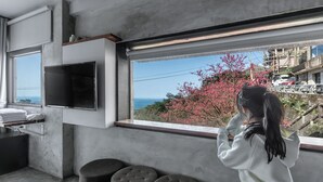 A modern room with a large window, a flat-screen TV mounted on the wall, a seating area with ottomans, and a view of the sea and pink flowering trees. - Jiufen The Ore Inn (New Taipei City)