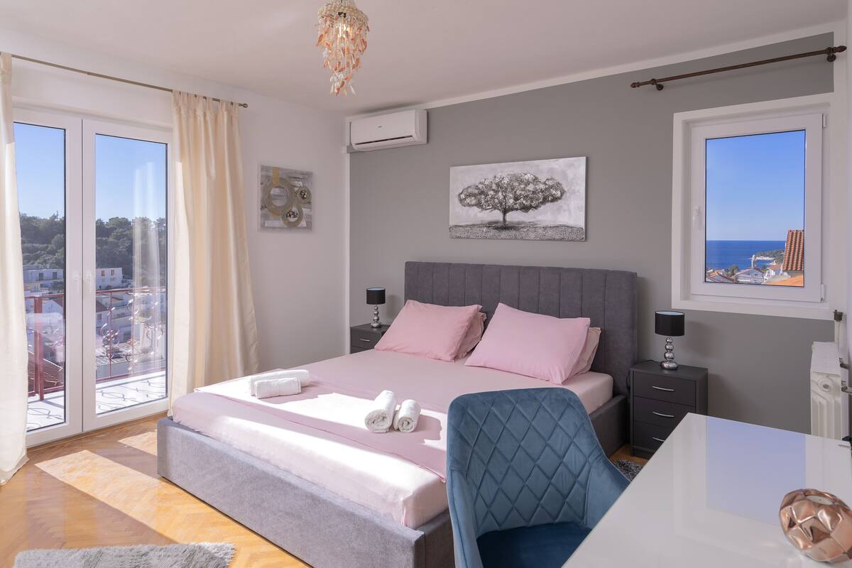 A bedroom with a bed, a desk, and a chair. There is a window with a view of the city and a balcony.