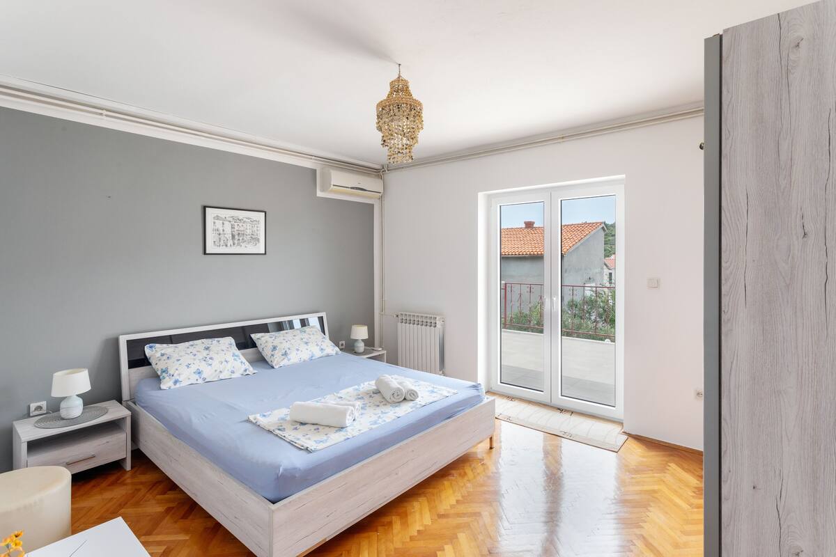 A bedroom with a large bed, a nightstand, a bedside table, a chandelier, a window with a view of buildings, and a radiator.