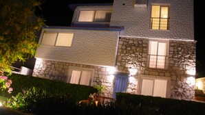 A two-story building with stone and stucco exterior, featuring multiple windows and a well-maintained hedge in front. - Tepe Hotel Alaçatı (Cesme)