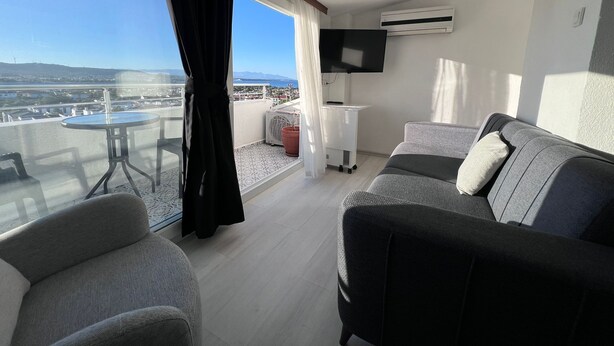 A balcony with a table and chairs, a sofa, a TV, and an air conditioning unit. - Tepe Hotel Alaçatı (Cesme)