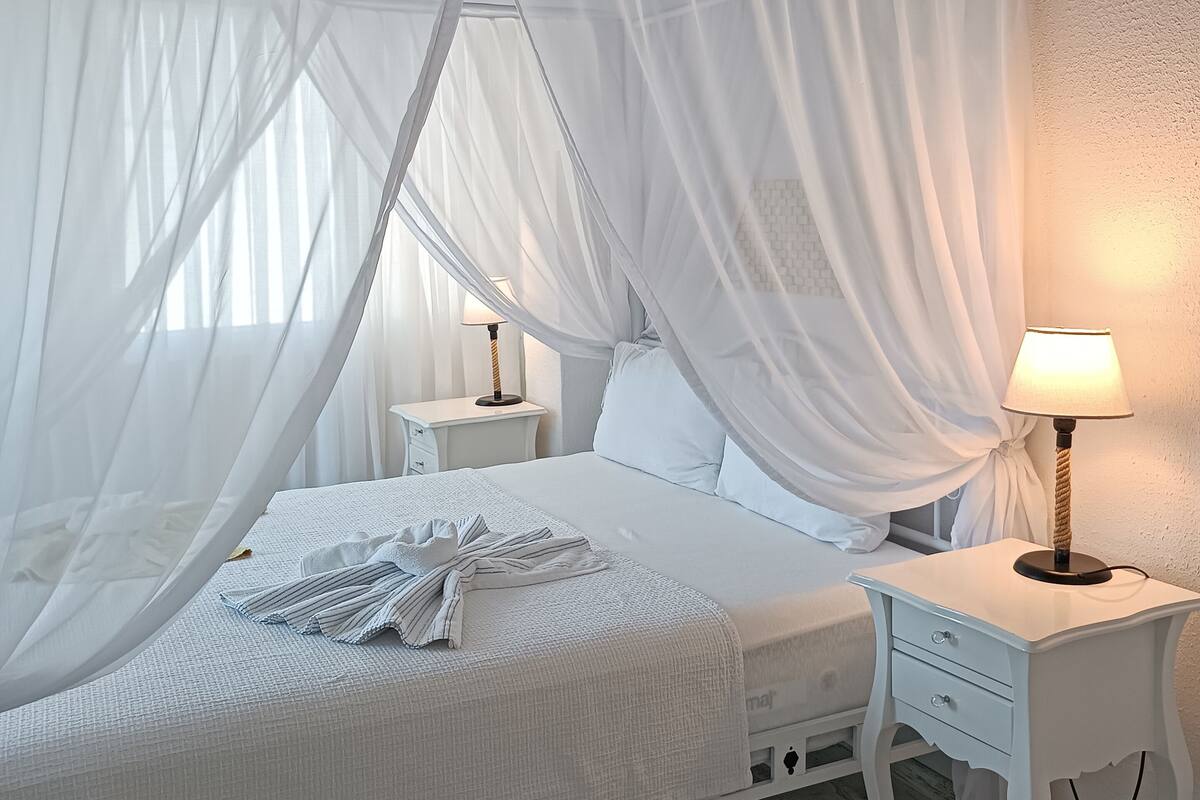 A bedroom with a canopy bed, white bedding, a bedside table with a lamp, and a nightstand.