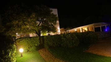 A nighttime view of a building with a well-maintained garden, a large tree, and a covered outdoor area with lights.