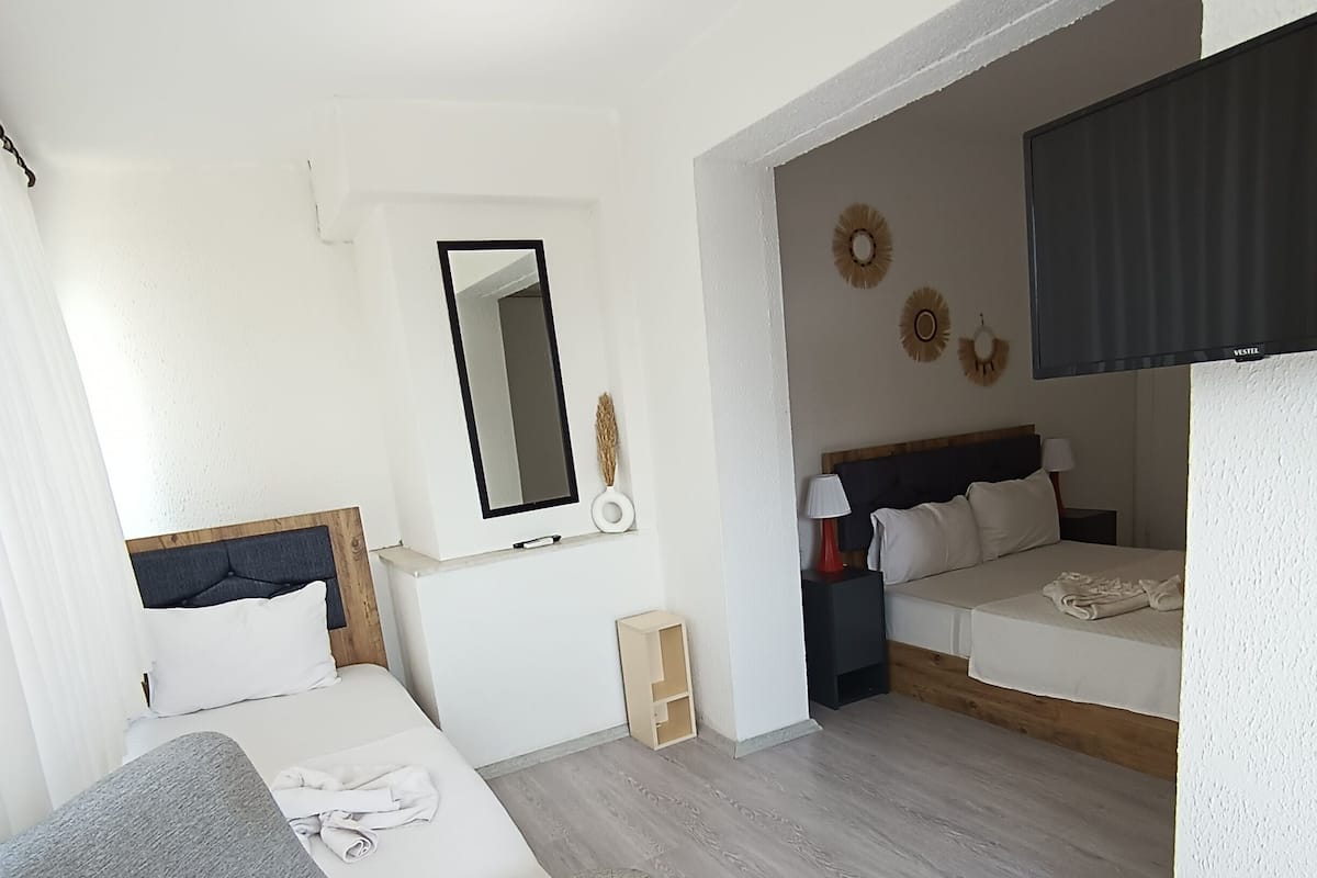 A modern hotel room with a grey sofa, a bed with white bedding, a small wooden nightstand, a wall-mounted TV, and a mirror.