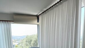 A modern living room with a grey sofa, a small armchair, and a white side table. A large window with sheer curtains offers a view of greenery and buildings. - Tepe Hotel Alaçatı (Cesme)