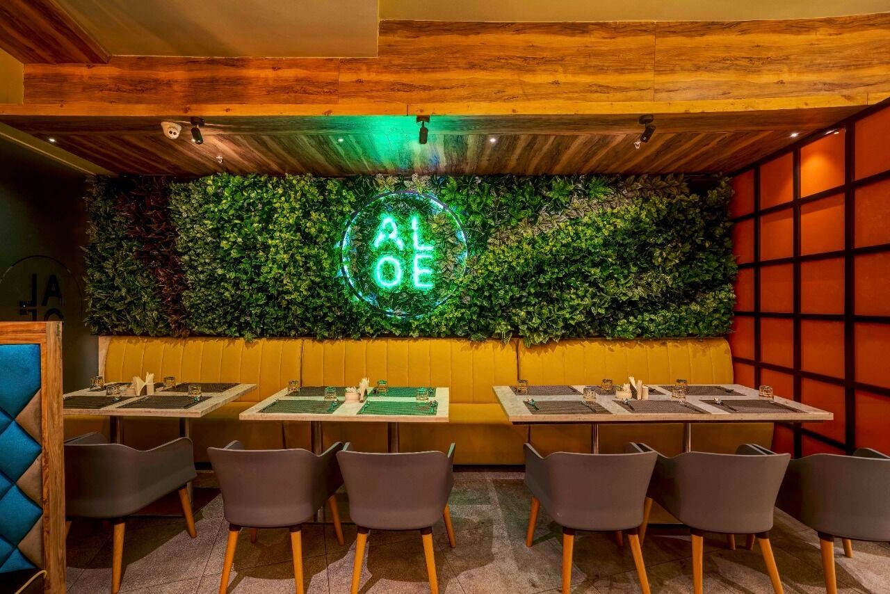 A restaurant interior with wooden ceiling beams, green foliage wall, neon 'ALOE' sign, and modern seating.
