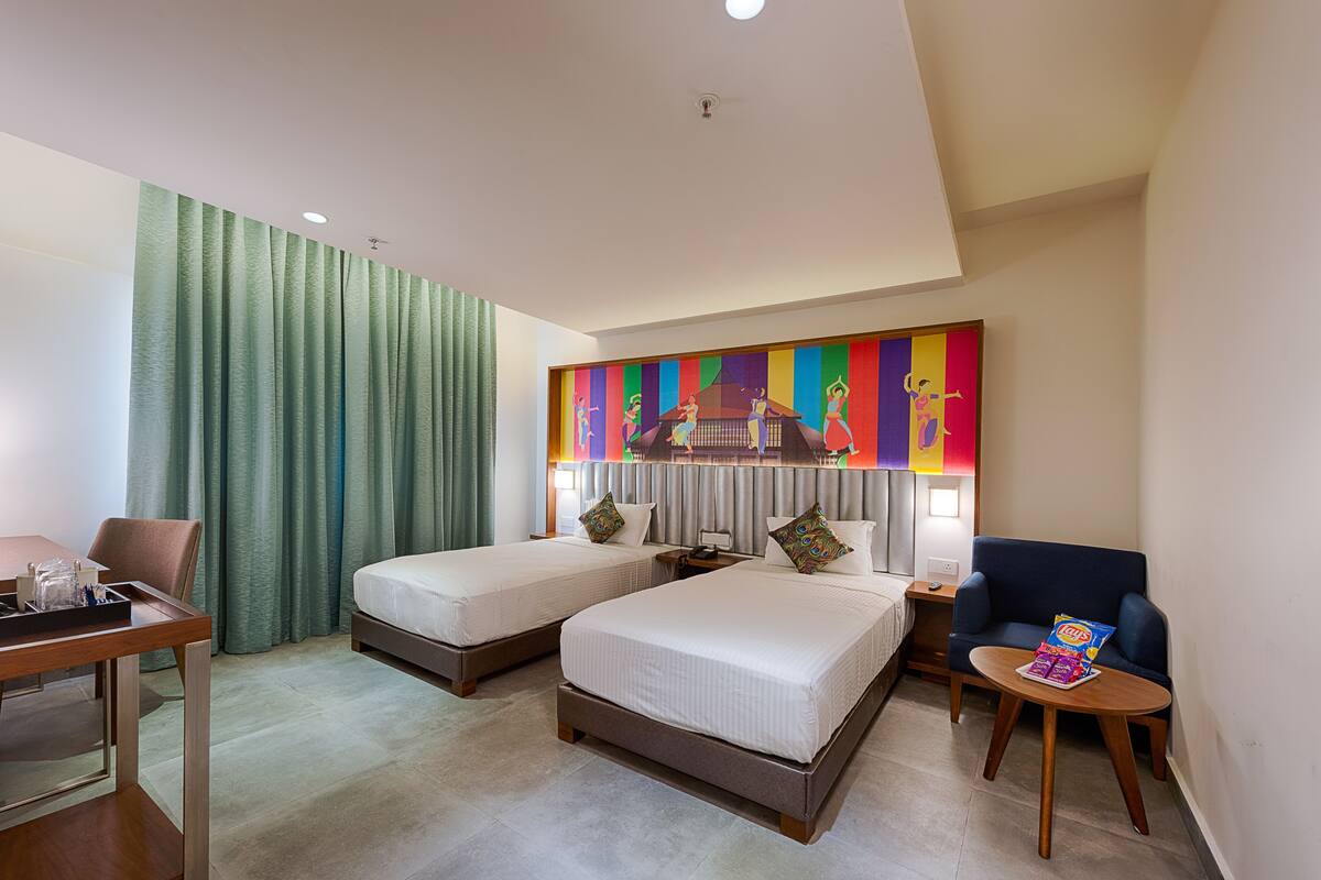 Une chambre d’hôtel avec deux lits, un bureau, une chaise, une petite table et un tableau coloré au mur.
