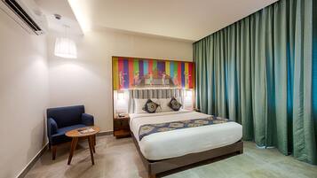 Una habitación de hotel con cama, una silla, una mesita pequeña, una mesita de noche y un colorido adorno en la pared.