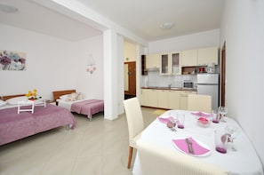 A modern hotel room with two beds, a dining table set for two, a kitchen area, and a small living space. - Apartments Roza (Seget)