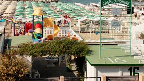 Um hotel à beira-mar chamado "BAGNI SUNSHINE", com telhado verde, área de recreação infantil e guarda-sóis de praia.