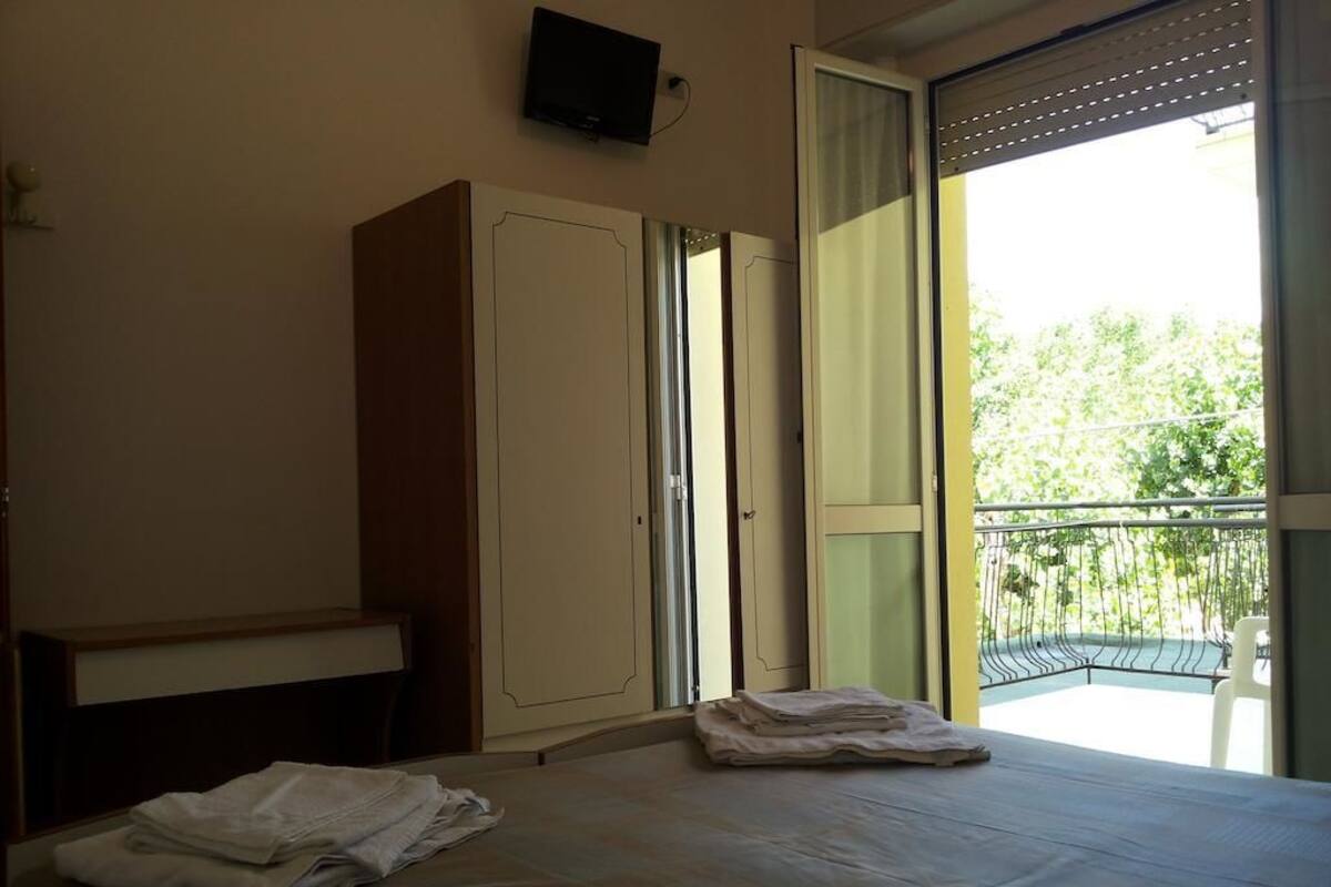 A bedroom with a bed, a wardrobe, a television, and a balcony with a view of greenery.