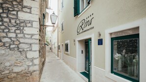 A narrow street with a building labeled "Riva de Lux Accommodation" featuring a stone facade, a green door, and a window with flowers.