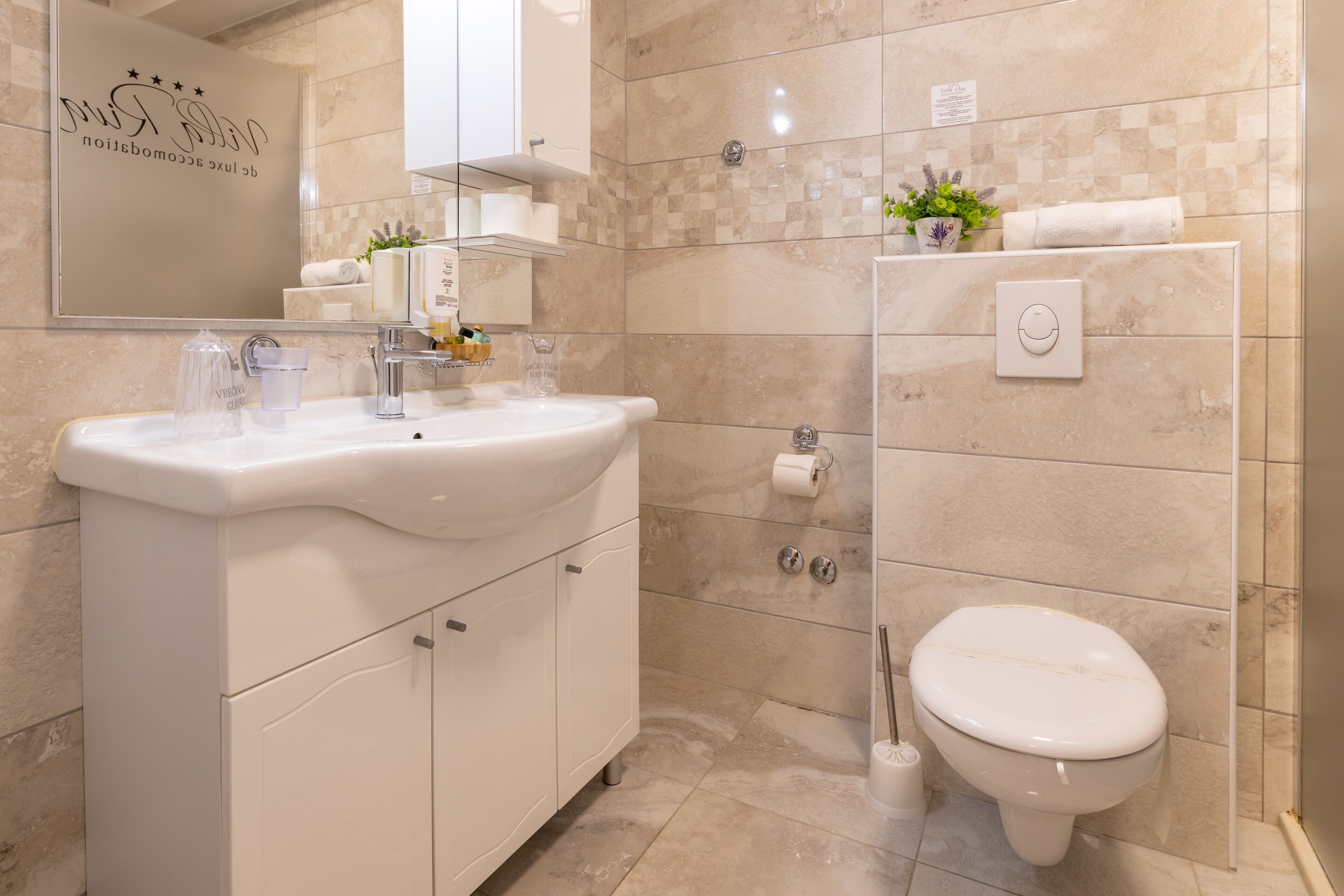 Un baño moderno con un lavamanos, espejo y retrete blancos. Hay artículos de aseo en el lavamanos y una planta en maceta en la pared.