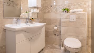 Un baño moderno con un lavamanos, espejo y retrete blancos. Hay artículos de aseo en el lavamanos y una planta en maceta en la pared.