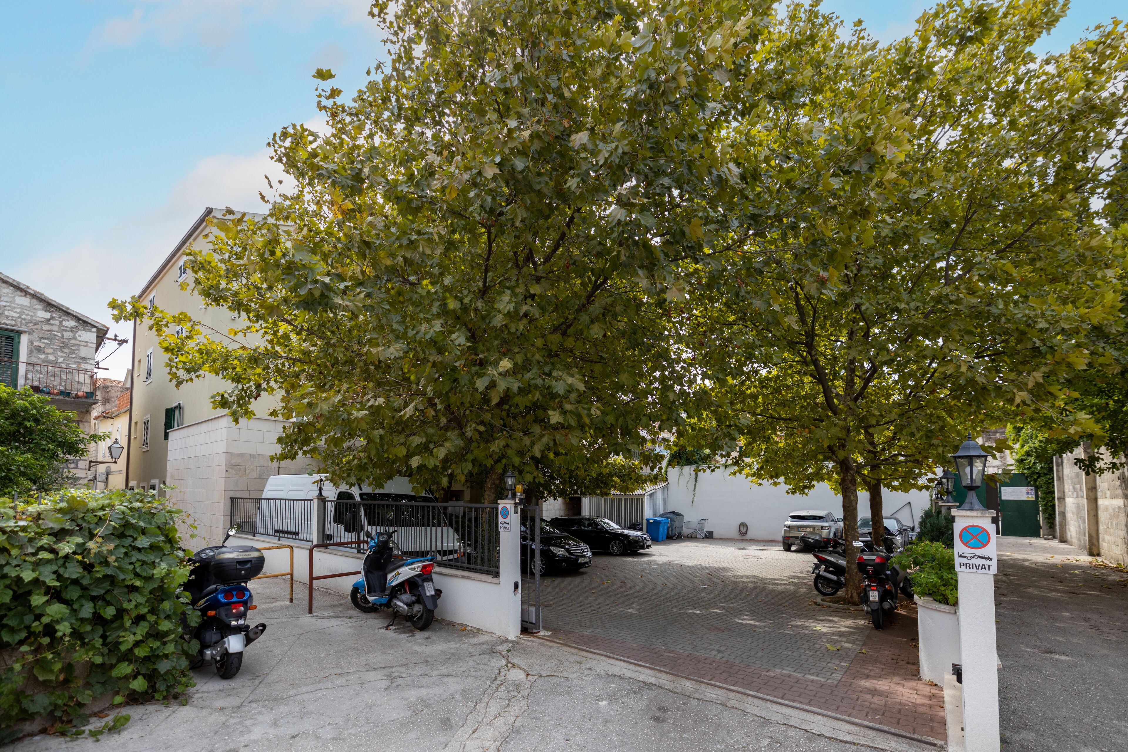 Un espace de stationnement clos, où se trouvent des motos et une voiture, le tout entouré d’immeubles et d’un arbre.