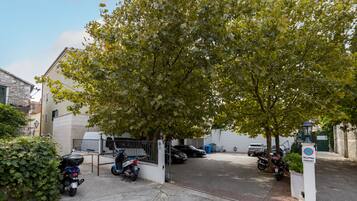 Un espace de stationnement clos, où se trouvent des motos et une voiture, le tout entouré d’immeubles et d’un arbre.
