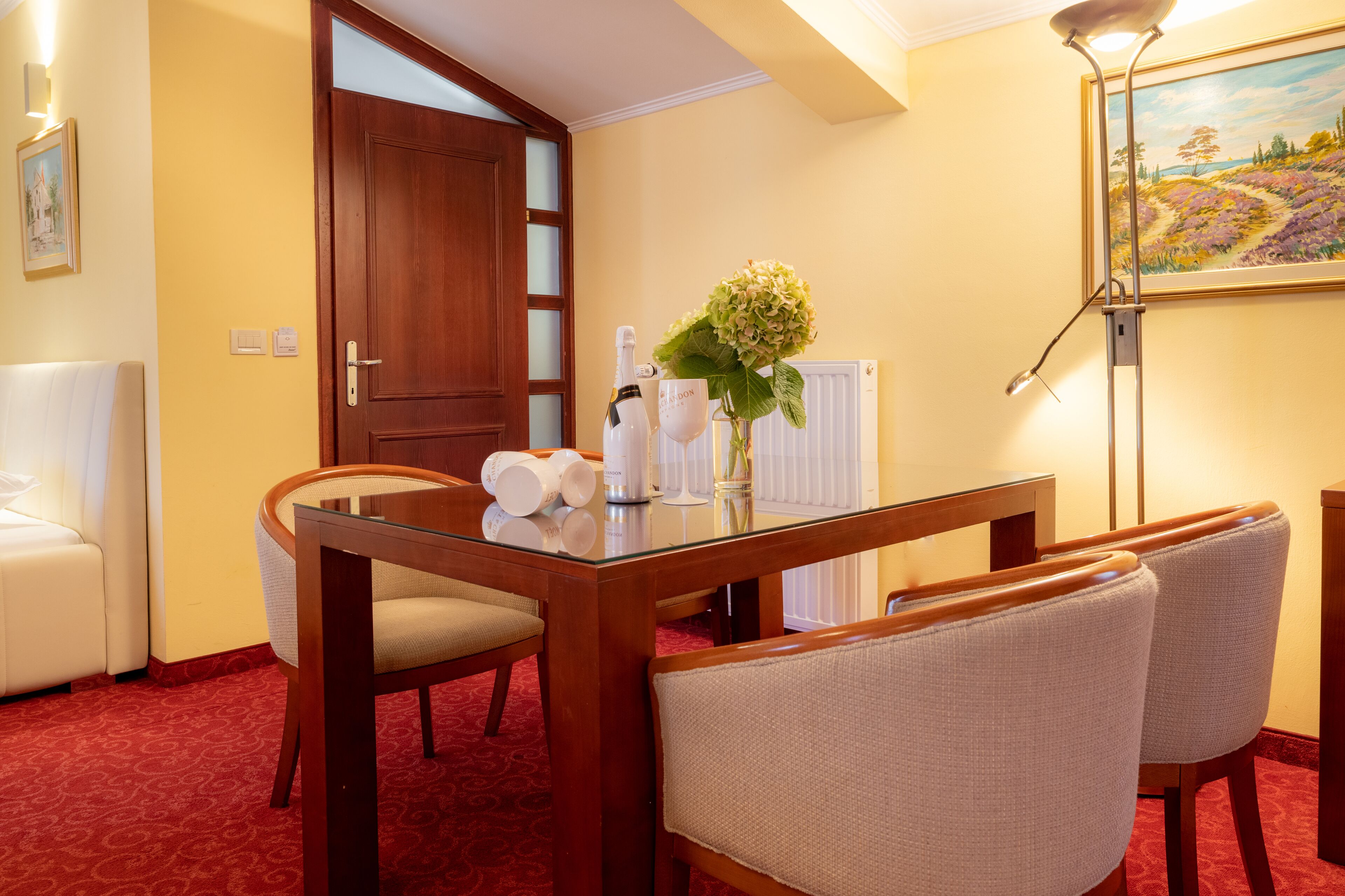 A hotel room with a wooden dining table, two upholstered chairs, a white sofa, a painting on the wall, and a door leading to another room.