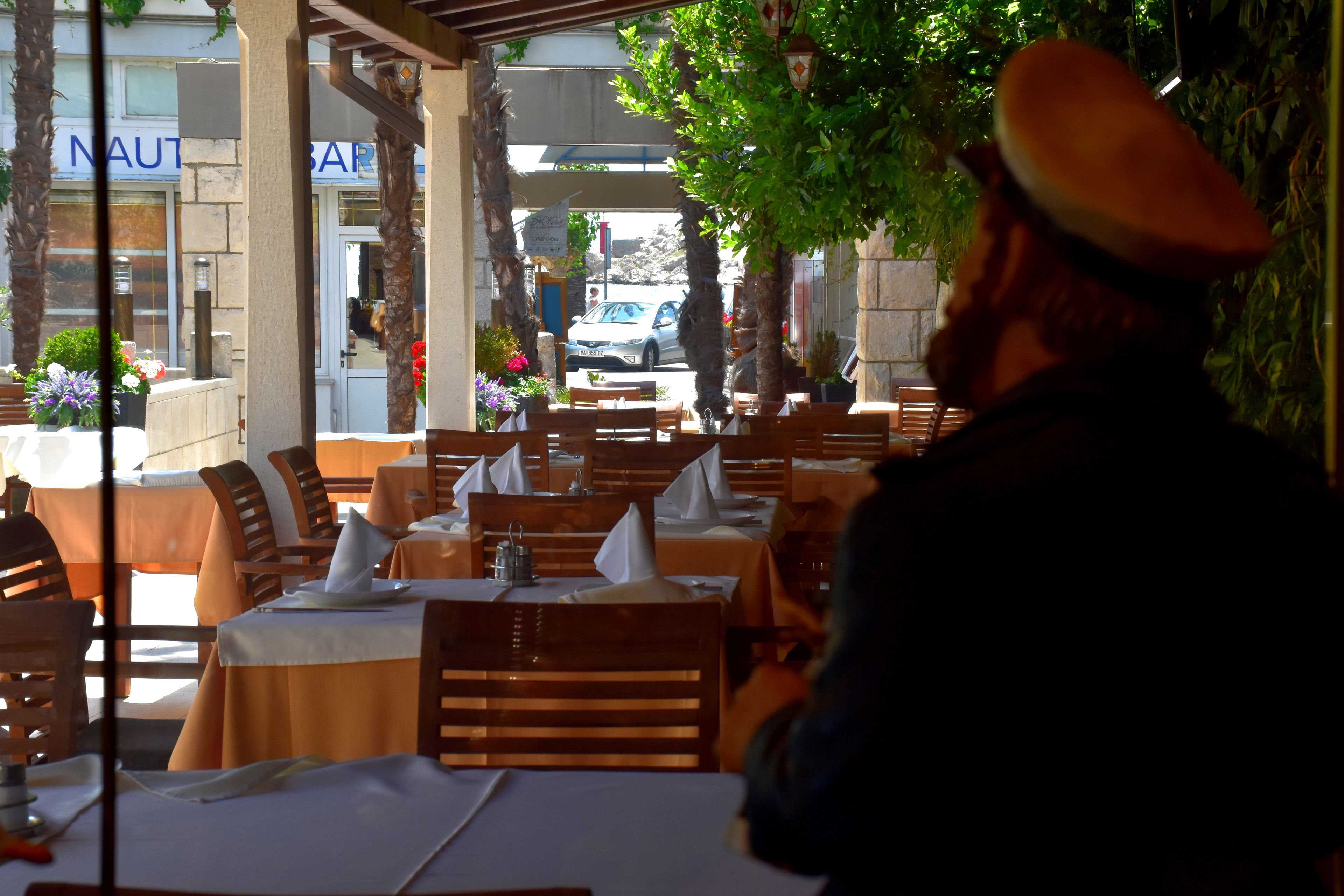An outdoor restaurant with tables and chairs, a tree, and a building with a sign in the background.