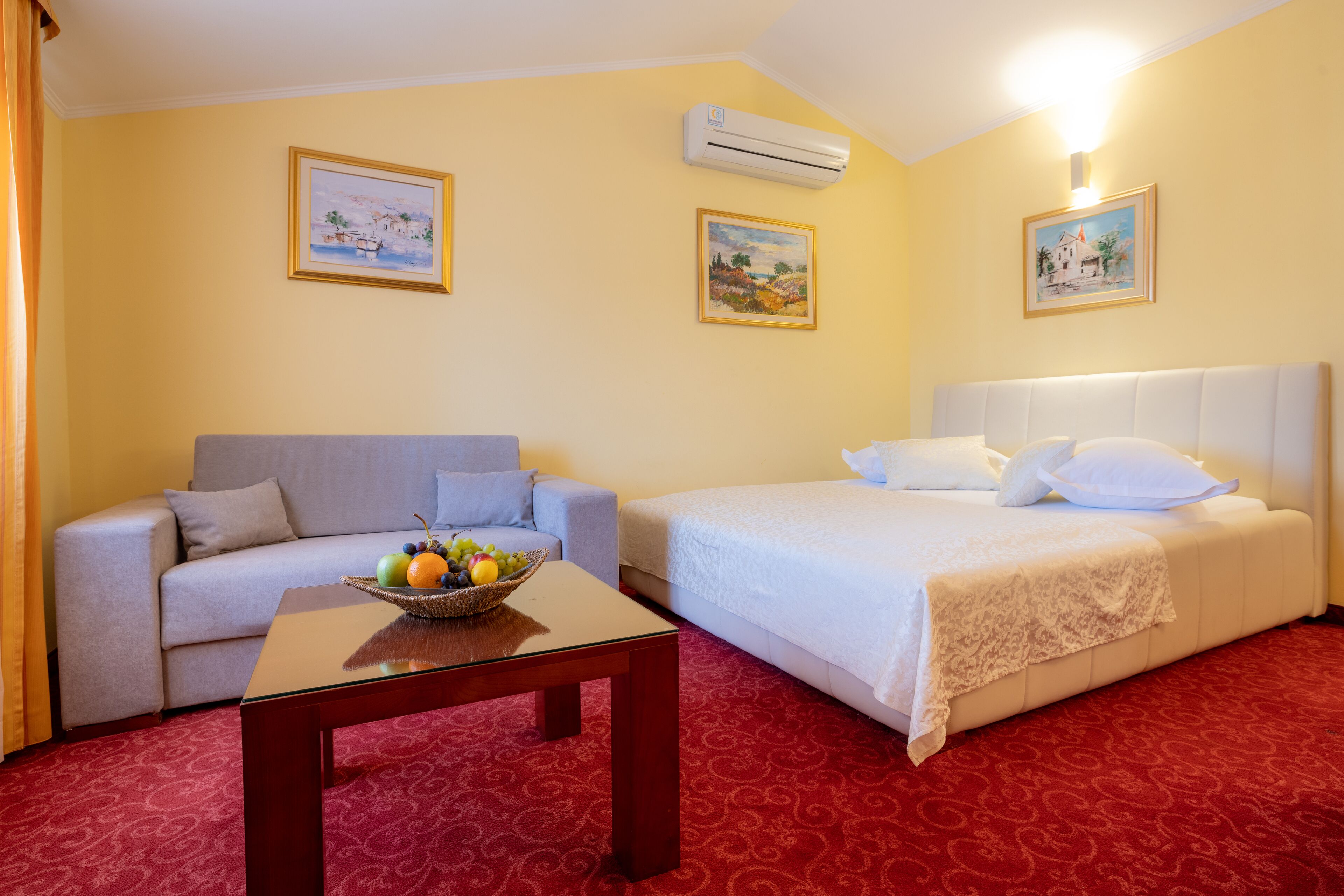 A hotel room with a bed, a sofa, a table with fruit, and two wall-mounted paintings.