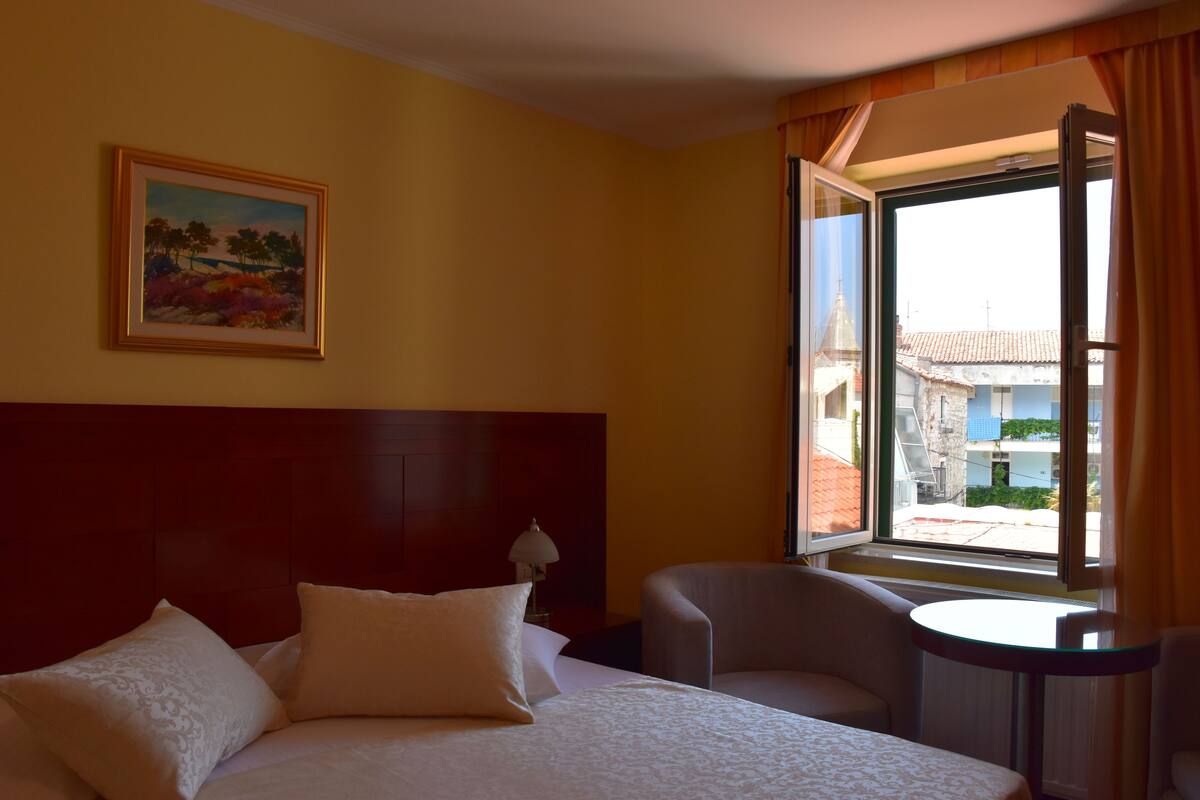 A hotel room with a bed, a chair, a small table, a painting on the wall, and a window with curtains.
