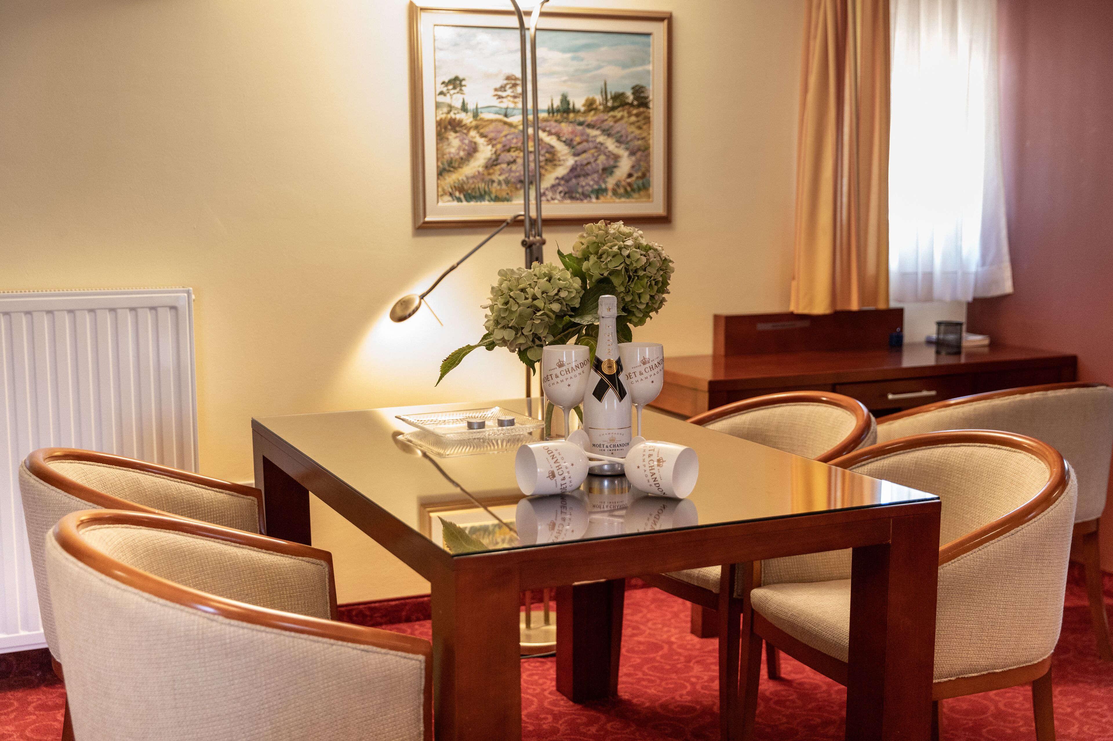 Un comedor de hotel con una mesa preparada para cuatro personas, un jarrón de flores y un cuadro en la pared.