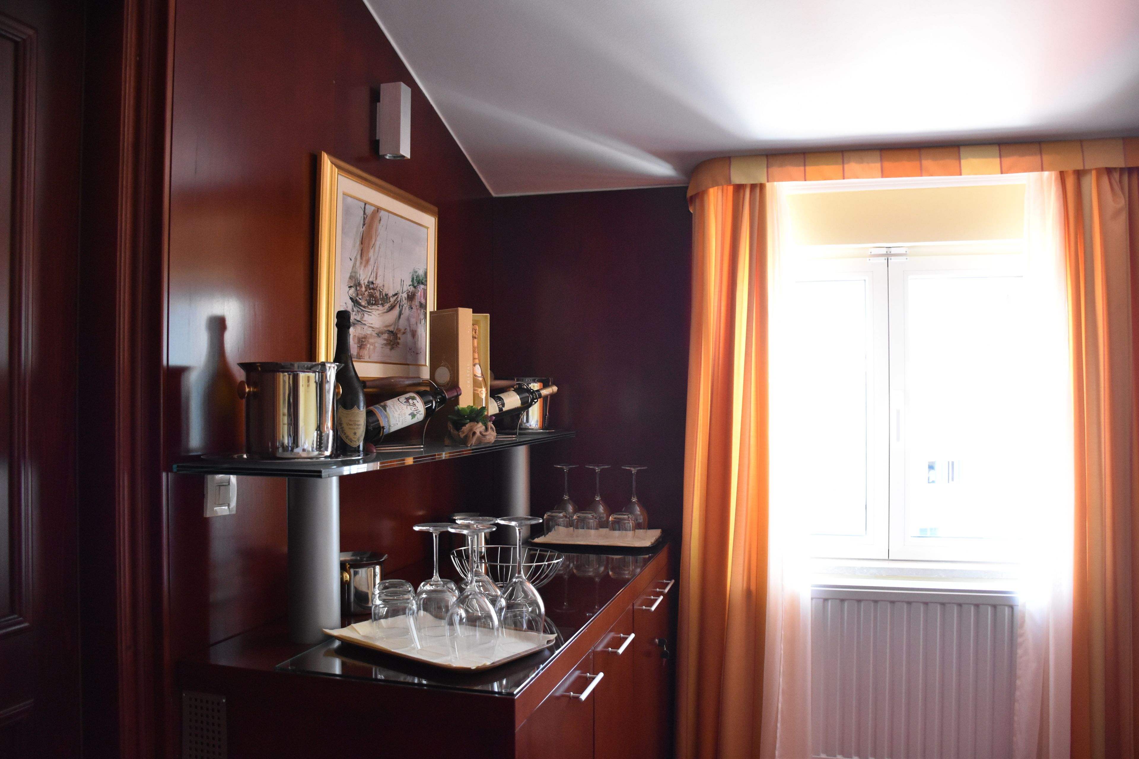 A bar area with wine glasses and bottles, a framed picture, and a window with curtains.