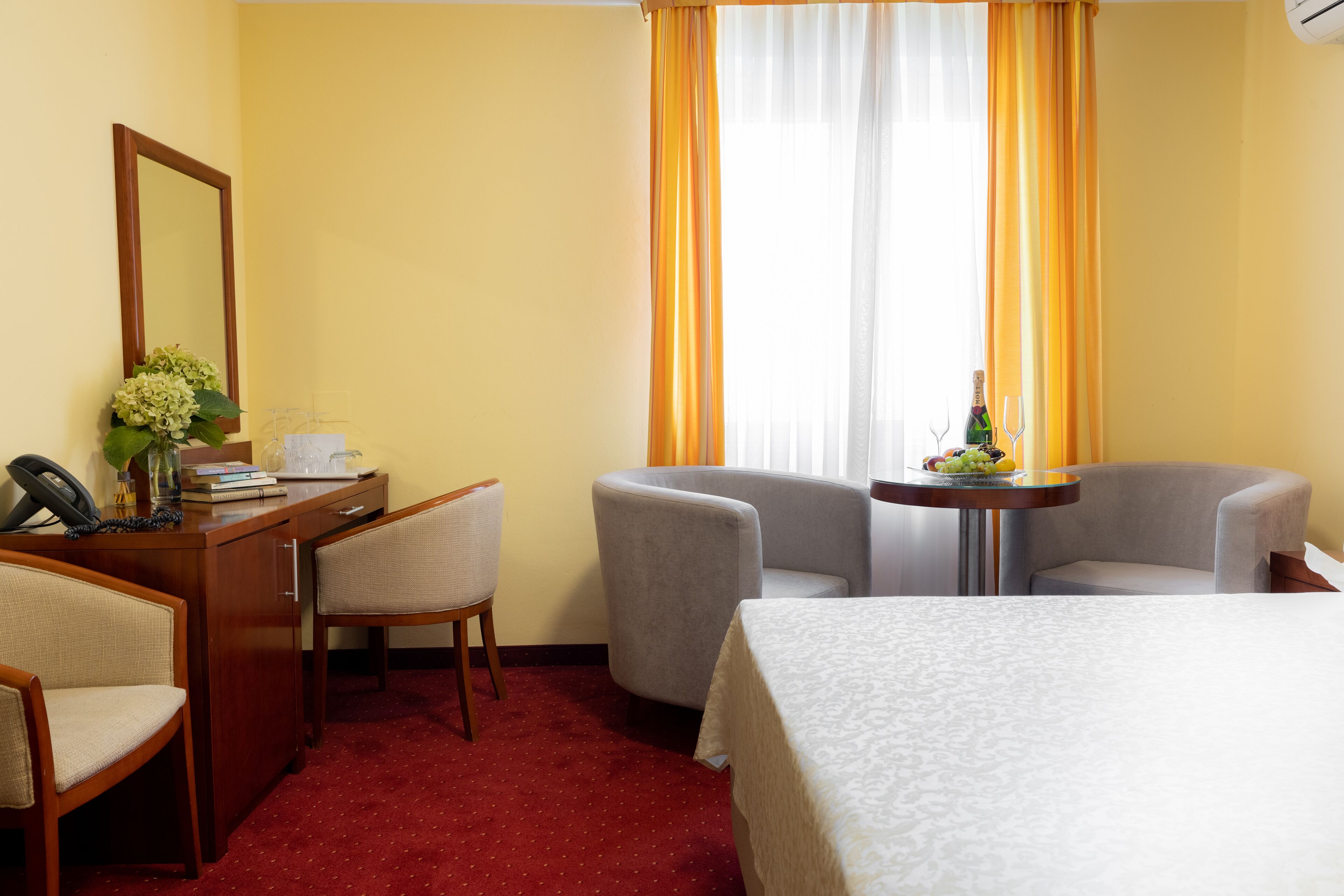 A hotel room with a bed, two armchairs, a desk with a telephone and a vase of flowers, a small table with a bottle and a glass, and a window with curtains.