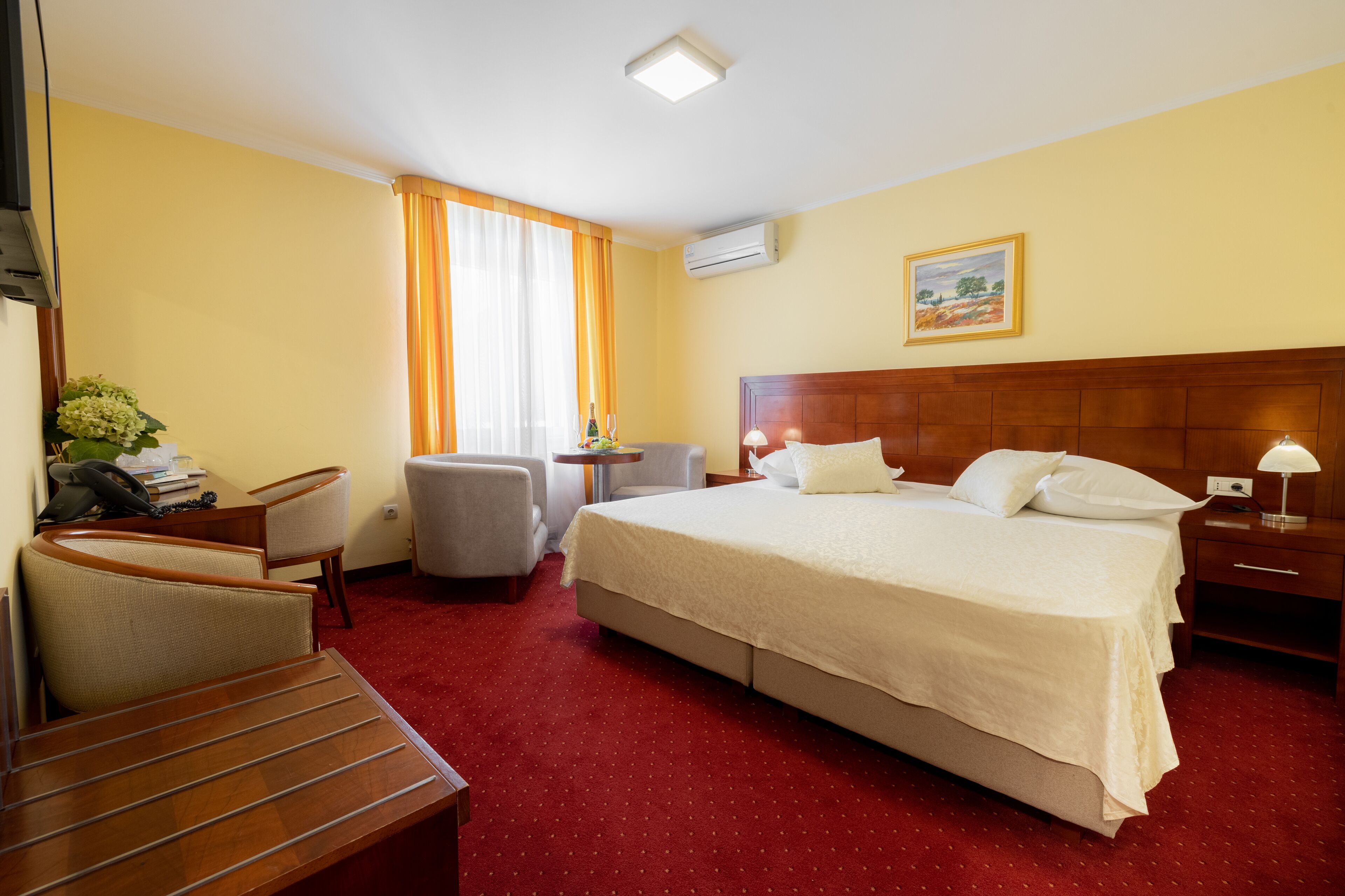 Habitación de hotel con cama, escritorio, silla, mesita pequeña y ventana con cortinas.