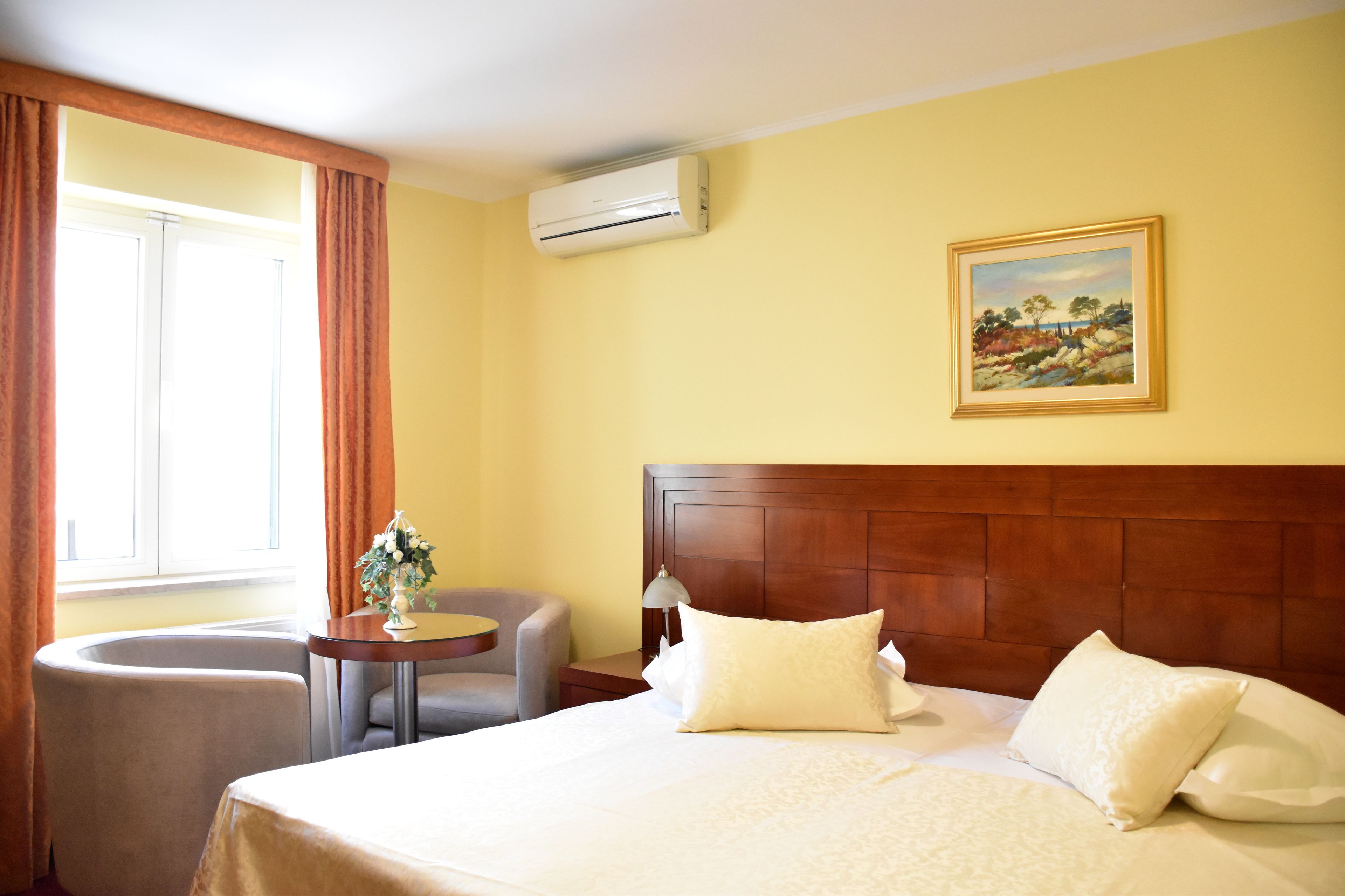 A hotel room with a bed, a chair, a small table, an air conditioner, and a painting on the wall.