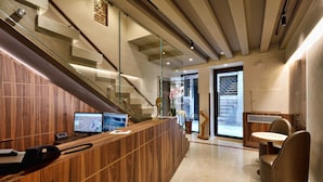 Un área de recepción con mostrador de madera, escalera de cristal y una zona de estar con una mesa redonda y sillas.