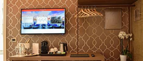 A hotel room with a wooden desk, a flat-screen TV displaying a cityscape, a coffee machine, wine glasses, and a potted orchid.