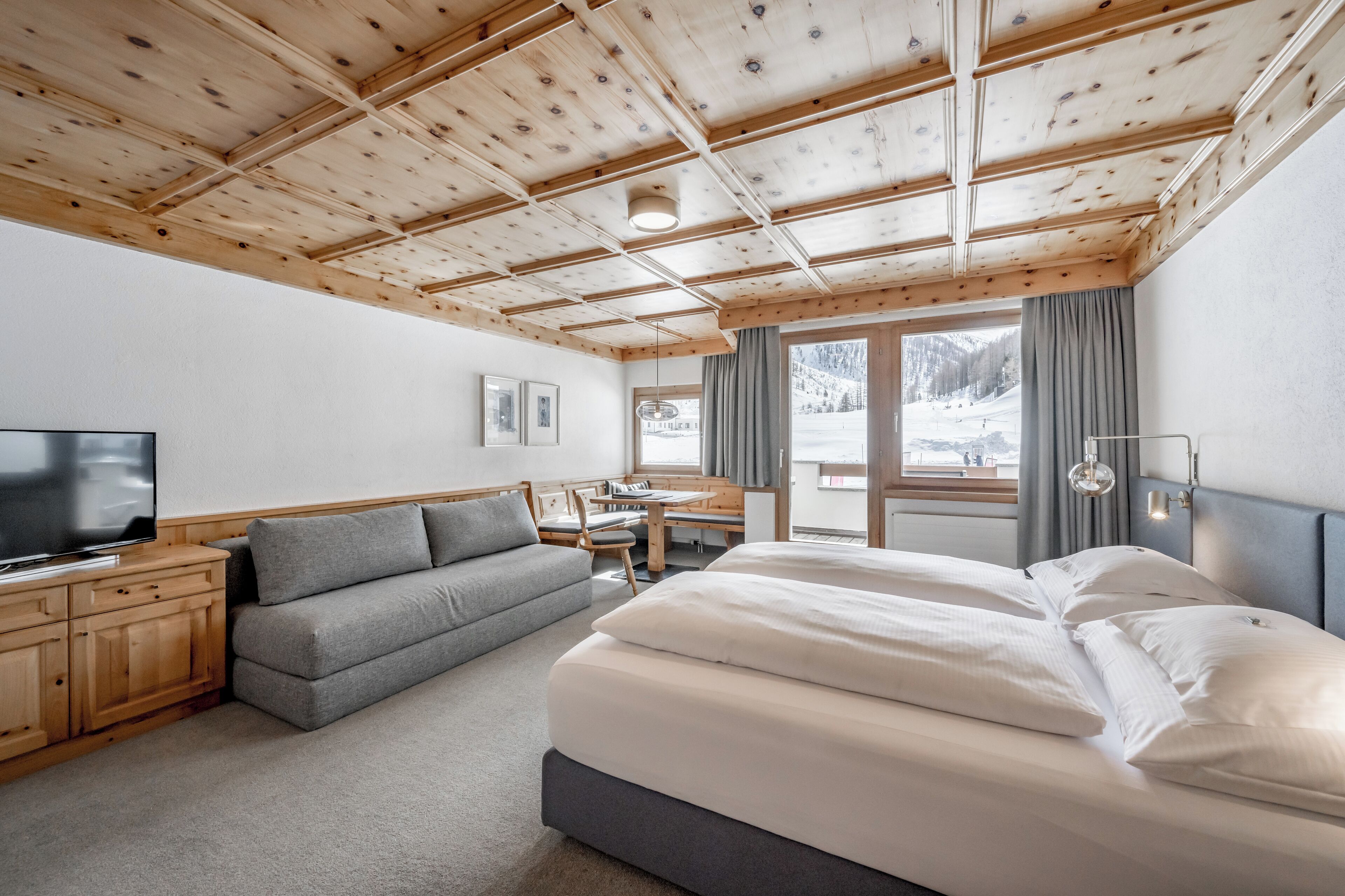A hotel room with a wooden ceiling, a bed, a sofa, a TV, and a window with a view of a snowy landscape.