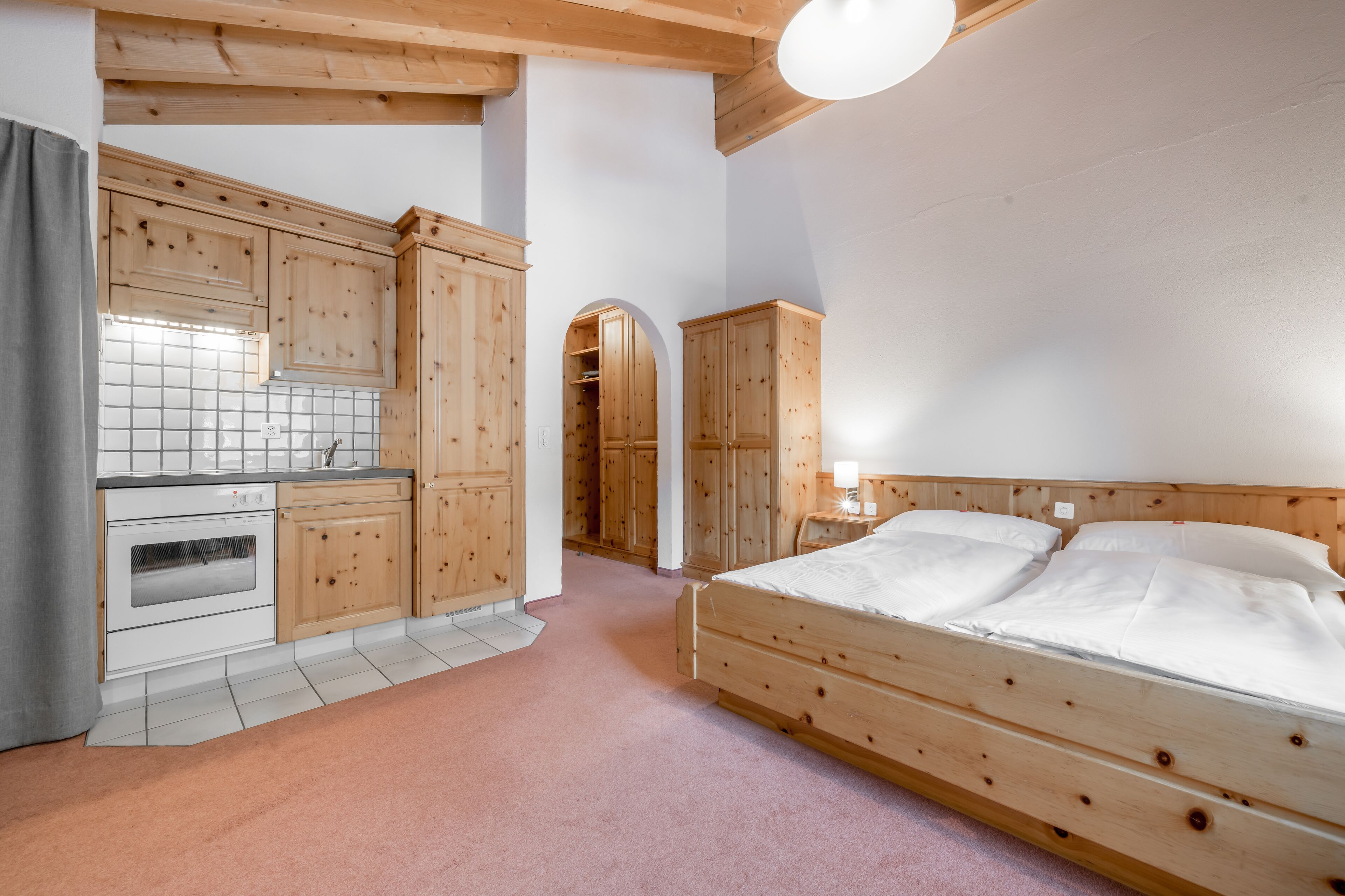 Un dormitorio con una cama de madera, una mesita de noche pequeña y un armario. Hay un armario de madera y una zona de cocina con estufa y armarios.